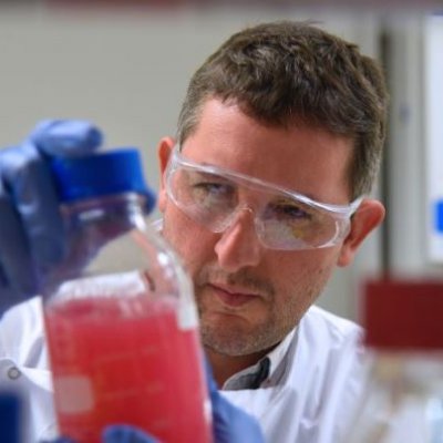 A male scientist in lab coat and goggles inspects a glass full of red liquid. 